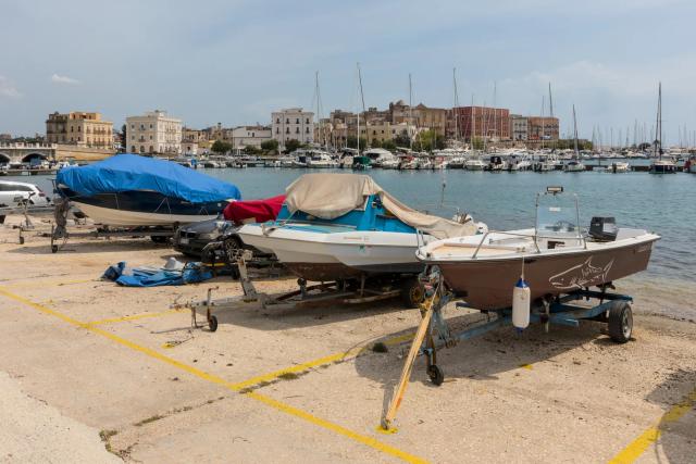Barche in rimessaggio su un piazzale davanti al mare, con la città vecchia di Taranto sullo sfondo