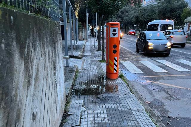 Un autovelox arancione a colonna sul marciapiede di una strada cittadina, con auto in transito e strisce pedonali