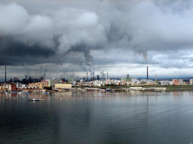 Le ciminiere dello stabilimento siderurgico di Taranto viste dal Mar Piccolo, con le case dei Tamburi e i filari dei mitili in primo piano