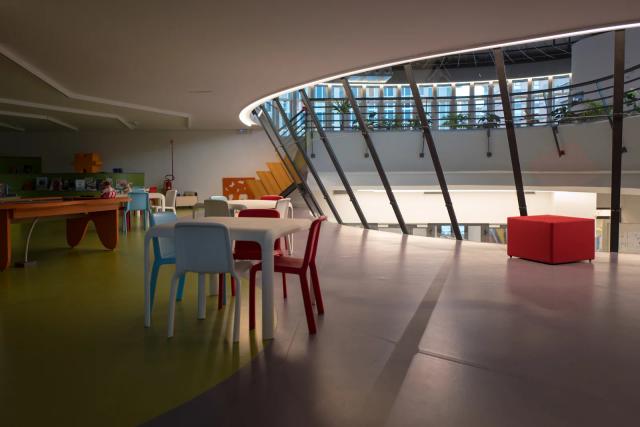 Una sala della Biblioteca civica Acclavio con tavolini e sedie colorate per bambini, scaffali bassi e una grande vetrata curva