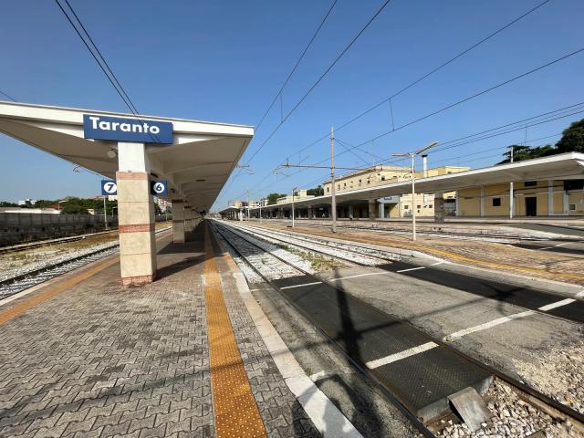 Un marciapiede della stazione di Taranto con il cartello blu del nome, la pensilina e i binari che si allontanano