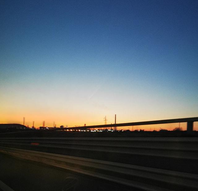 Le ciminiere dello stabilimento ex Ilva di Taranto in controluce al tramonto, viste dalla strada, sotto un cielo che sfuma dall’arancio al blu
