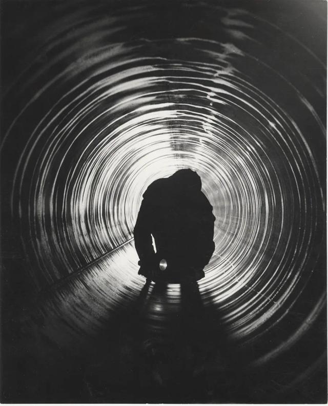 Foto in bianco e nero del 1963: un operaio Italsider, chino con una torcia, controlla l’interno di un grande tubo d’acciaio